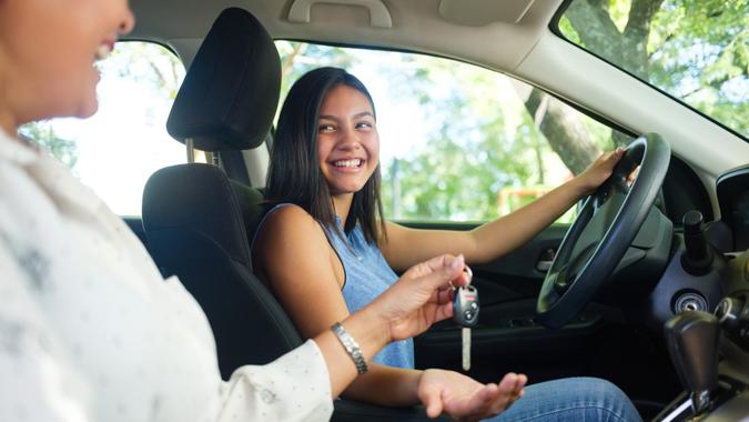 Side view photo of a teenage girl in a car, her mother sitting next to her and handing her the keys to drive.