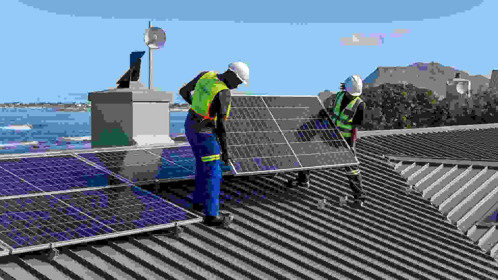 Two African men install solar panels neatly on a residential roof of a house near the ocean.