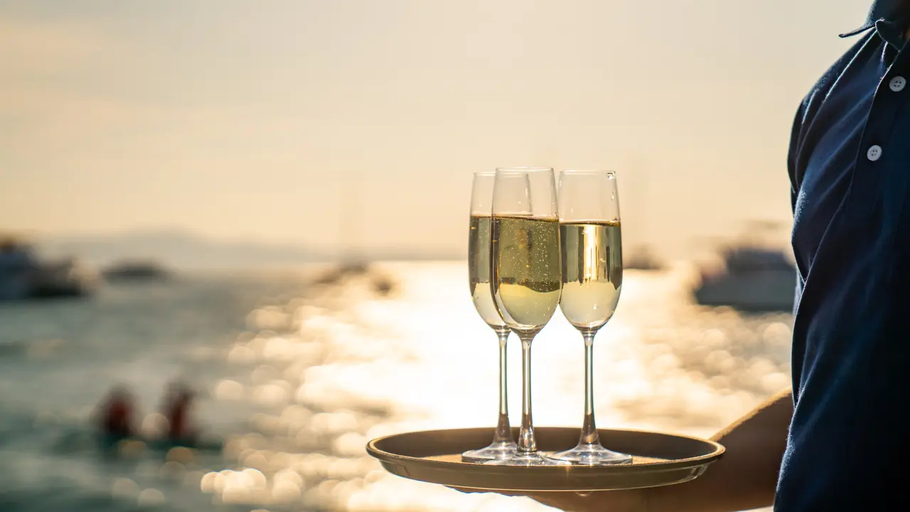 Asian man waiter holding champagne glass on the tray for serving to passenger tourist while luxury catamaran boat sailing in the ocean at summer sunset.