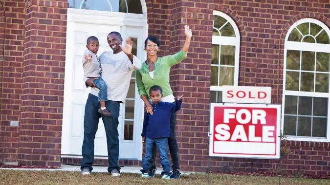 Happy African American family with two young children (4-5 years) waving in front of house with 'Sold' sign.