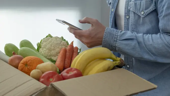 MAN HOLDING SMARTPHONE RECEIVING PACKAGE OF FRUITS AND VEGETABLES AT HOME. ONLINE PURCHASE ORDERS OF ECOLOGICAL PRODUCTS. VEGAN AND VEGETARIAN FOOD CONCEPT. stock photo