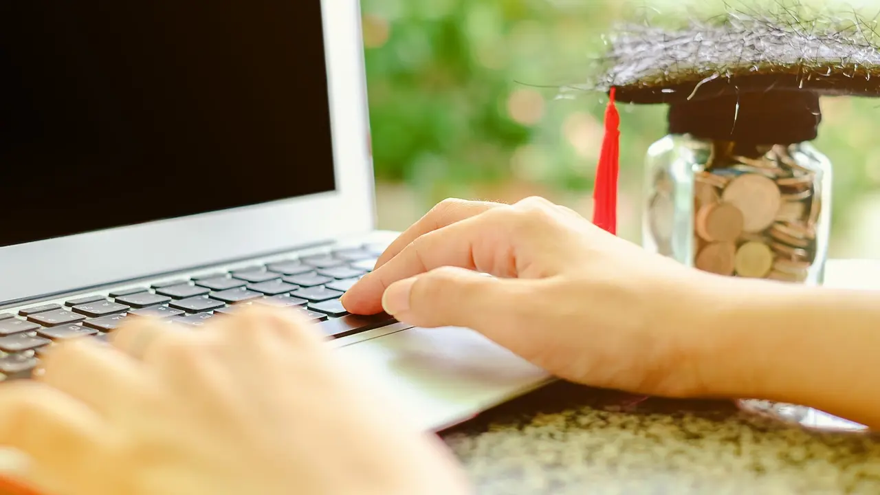 Hand typing on computer laptop with square academic cap and the glass jar of coin on blurred natural green background for education, finance and saving money concept stock photo