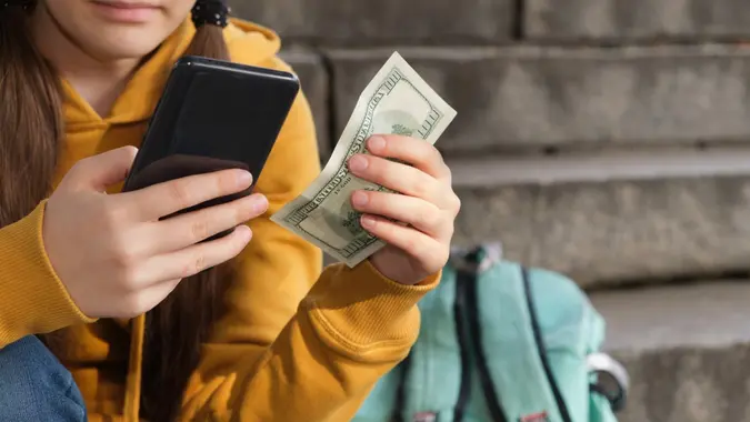The teenager holds dollar bills and looks at the smartphone screen.  stock photos