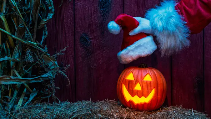 Santa's gloved hand placing a Santa Hat on a carved glowing happy Jack O' Lantern looking at the camera sitting on a bale of straw with dried corn stalks and a weather red barn wall in the background at night under a blue moon light.