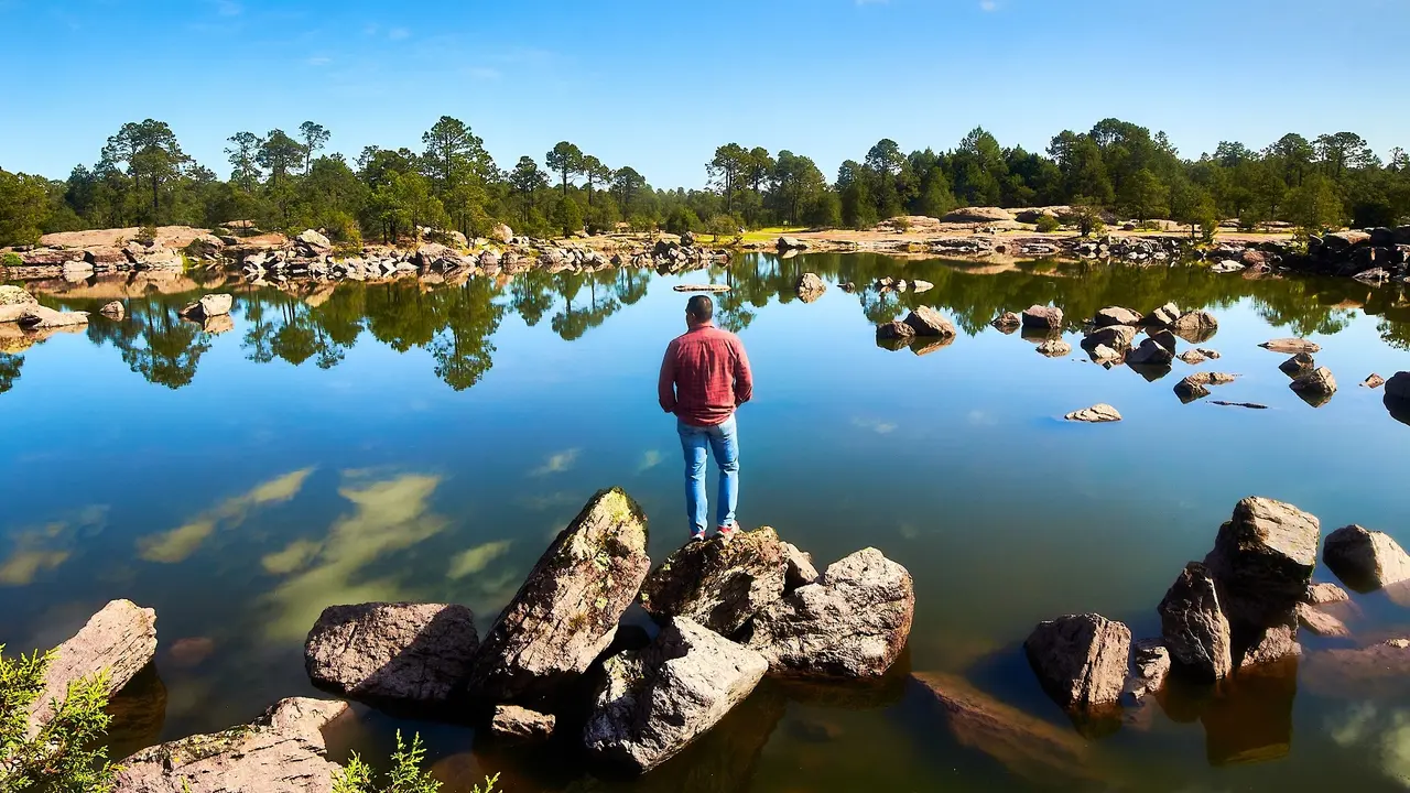 Man standing on a rock in front of a reflecting lake with a forest in the background on a sunny day in Mexiquillo natural park in Durango stock photo