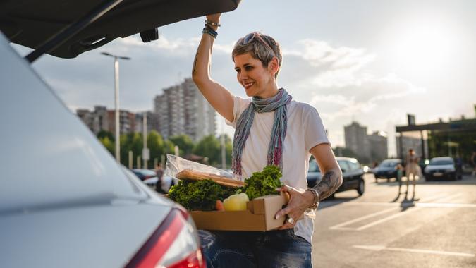 One woman mature caucasian female standing by the back trunk of her car on the parking lot of the supermarket shopping mall or grocery store with vegetables food in box putting them in the vehicle stock photo