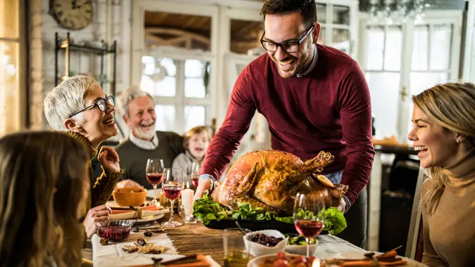 Happy man serving roasted turkey to his family during Thanksgiving dinner at dining table.