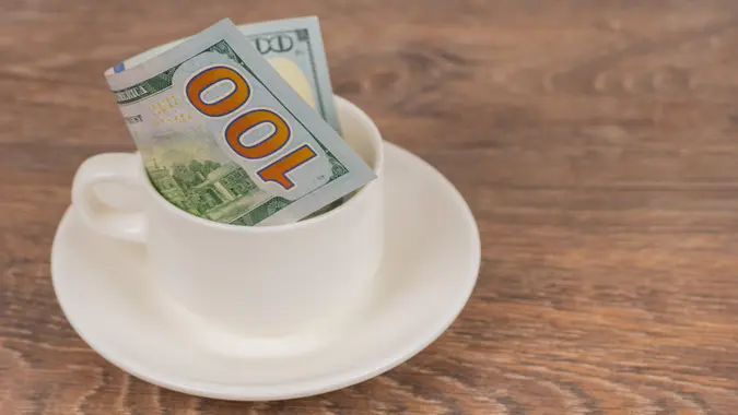100 dollars banknote in white coffee cup on wooden table, tips or expensive service concept.