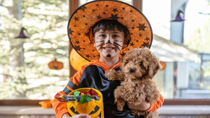 Cute Child In Costume With His Dog Holding Bowl Full Of Candies On Halloween At The Front Door.