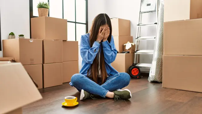A young woman sits on the floor and holds her hands over her face.