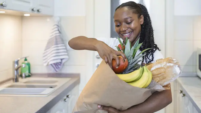 Healthy positive happy woman holding a paper shopping bag full of fruit and vegetables. Young woman holding grocery shopping bag with vegetables Standing in the kitchen stock photo