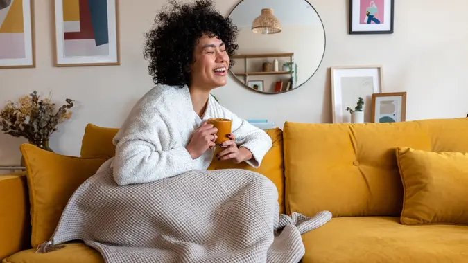 Happy African american woman enjoying quiet time at home laughing, drinking morning coffee sitting on sofa. Copy space stock photo