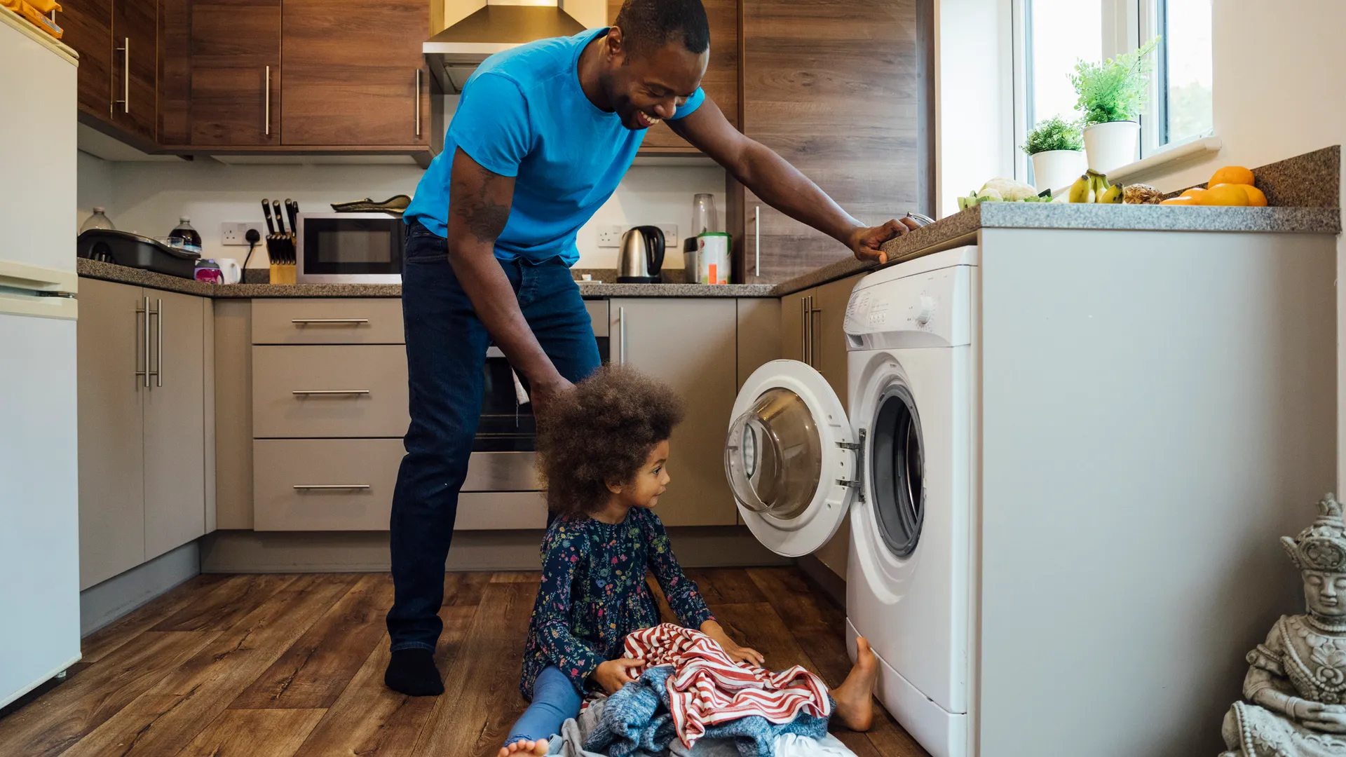 一个混血女孩坐在厨房的地板上，帮她父亲把洗好的衣服放进洗衣机。