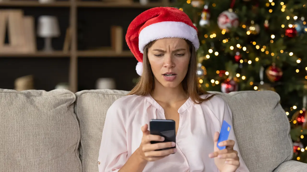 Worried concerned girl in Christmas Santa hat having problems with payment by credit card online for New Year purchases, looking at smartphone screen with puzzled face.