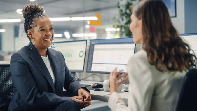 Two Female Colleagues Fondly Talk to Each Other, Laugh and Smile while Working on Computers in Diverse Modern Business Office.