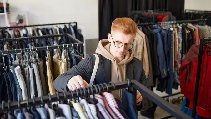 High angle portrait of red haired young man looking at clothes while shopping sustainably in thrift store.
