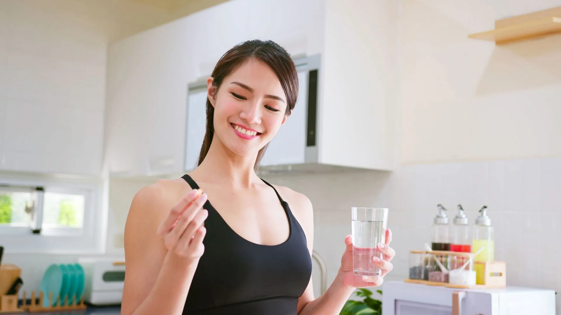 asian fitness woman taking nutrient supplement or medicine by water in kitchen at home.