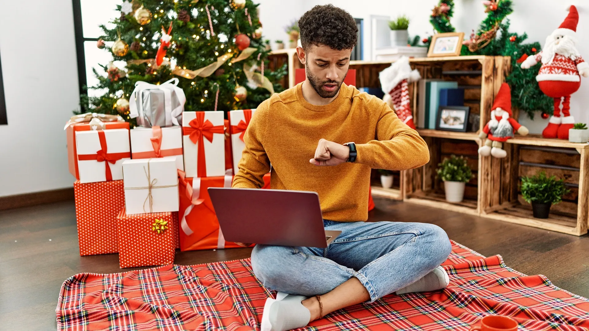 Arab young man using laptop sitting by christmas tree looking at the watch time worried, afraid of getting late stock photo