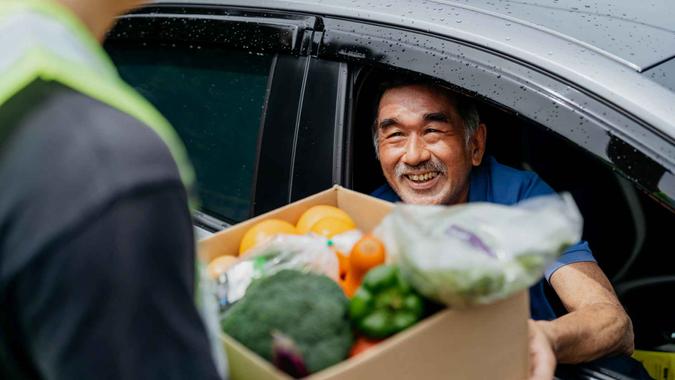 Image of an Asian Chinese senior man receiving a box of free food and groceries from volunteer at a drive through food bank.