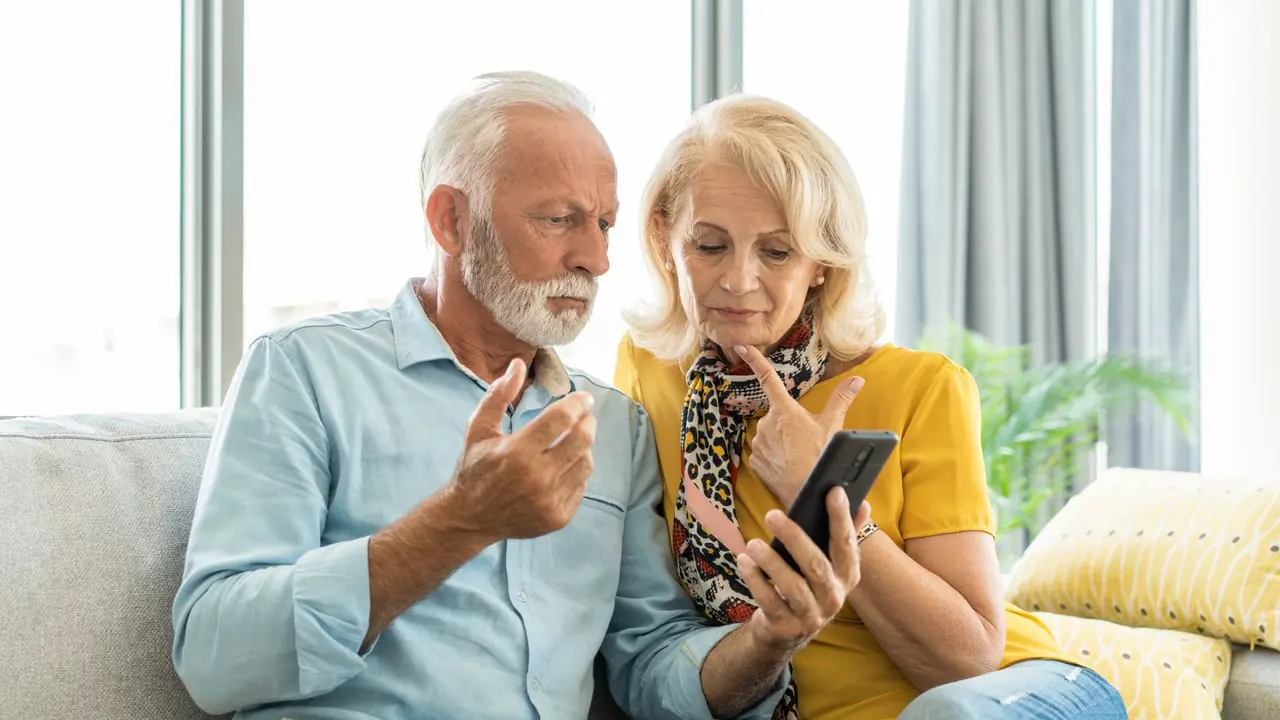 Worried senior couple using phone while sitting on sofa at home.