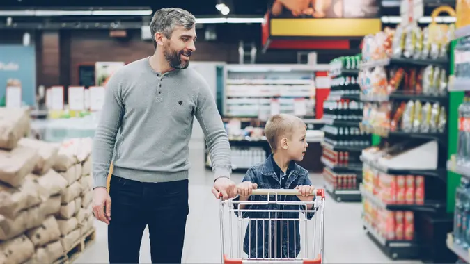 young family father and son walking through large food store with trolley looking around and talking.