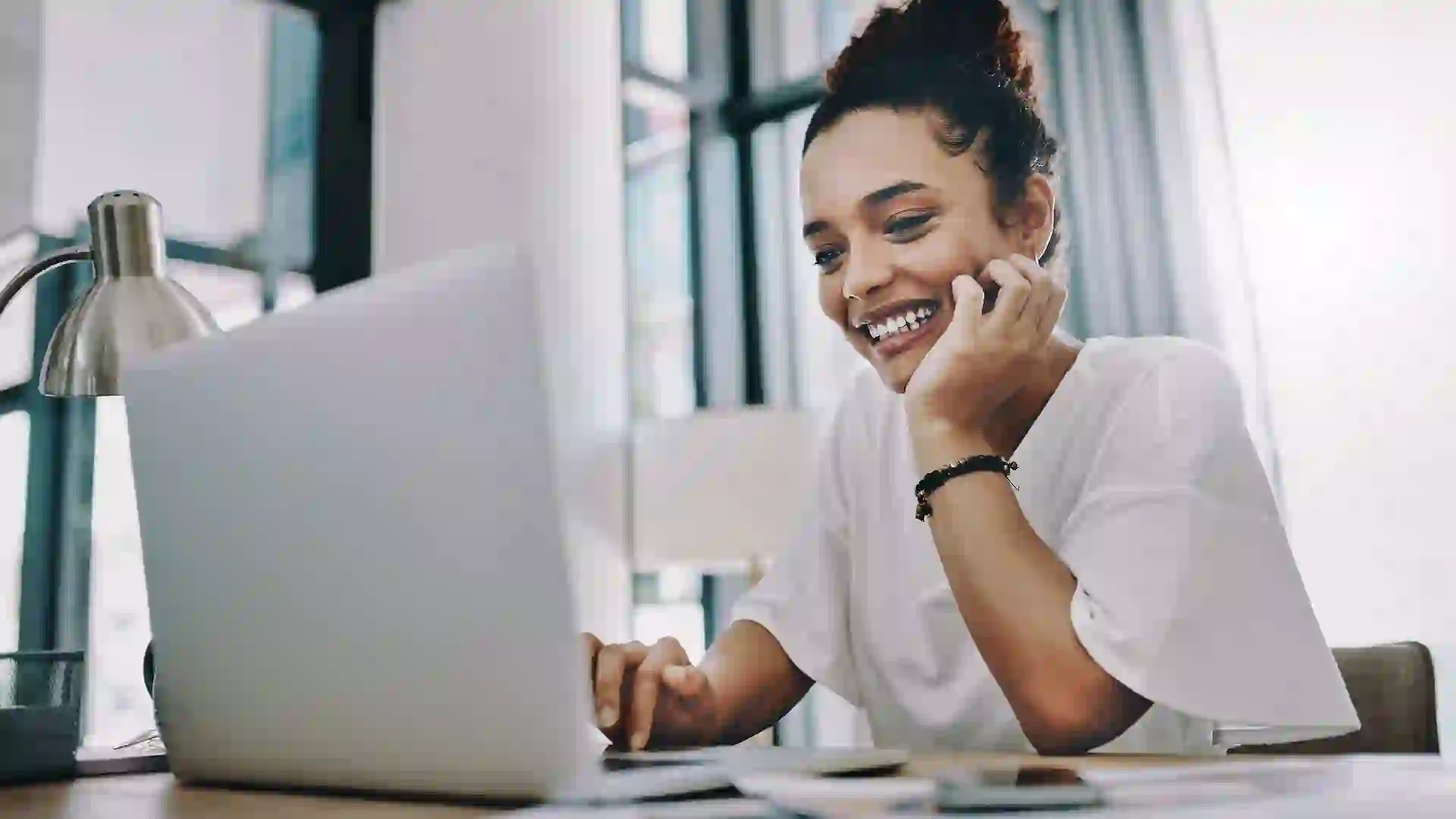 Happy woman smiling and sitting in front of windows at her desk while looking at an open laptop