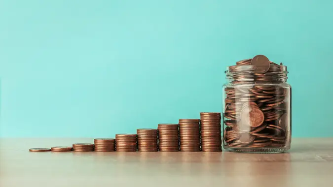 Stock photo of an ascending staircase of coins on a blue background with a jar full of coins.