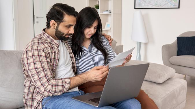 Young indian couple reading paper bills paying loan bank debt online together on computer, calculating taxes, income, making payments, planning family budget money finances using laptop at home.