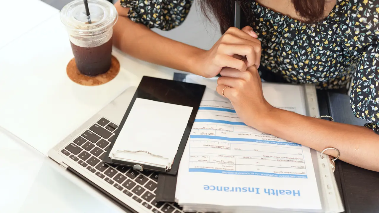 Person uses a calculator and laptop while reviewing financial documents at a wooden desk with coffee and a notebook nearby.