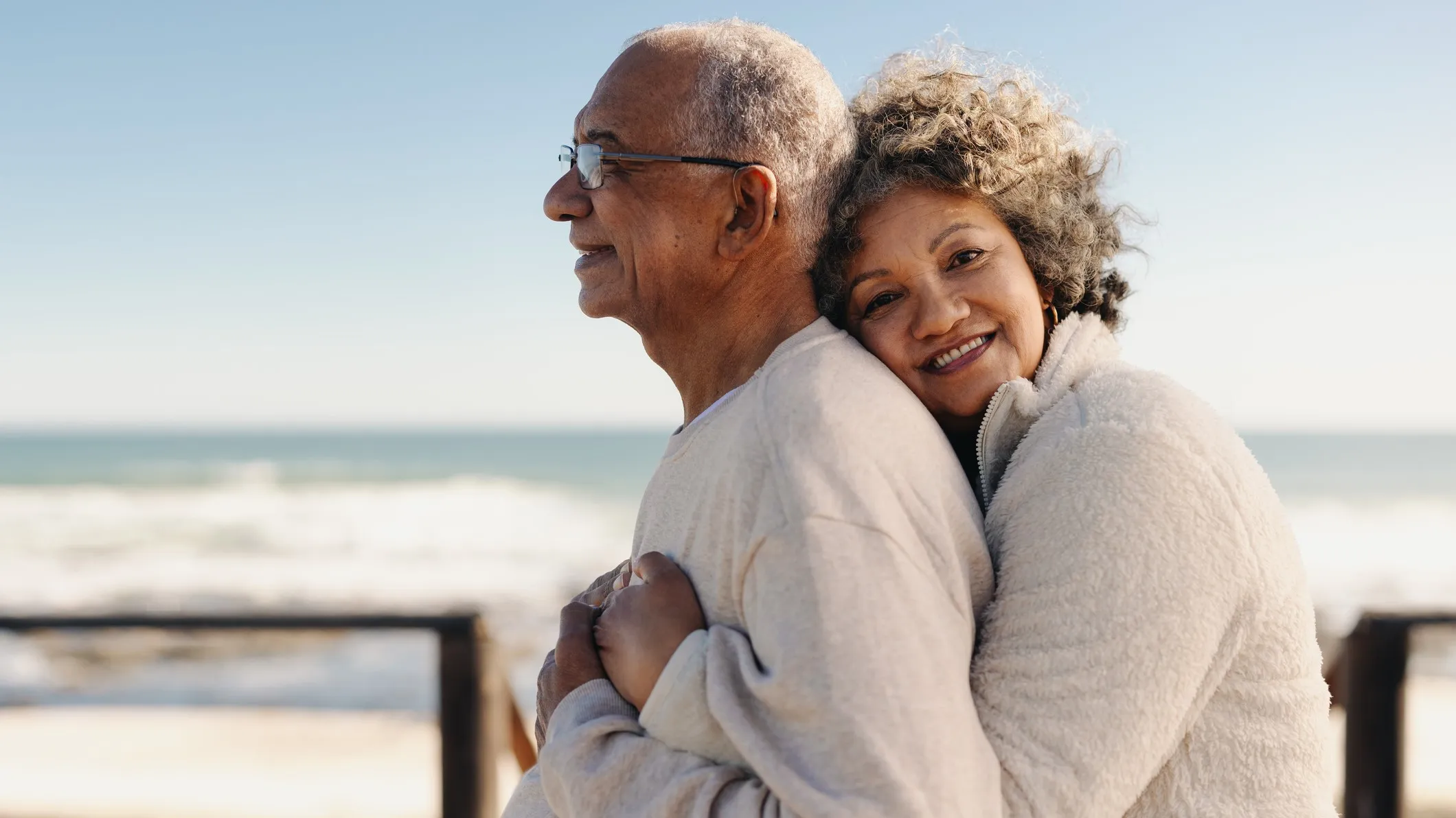 Romantic senior woman smiling at the camera while embracing her husband by the ocean.