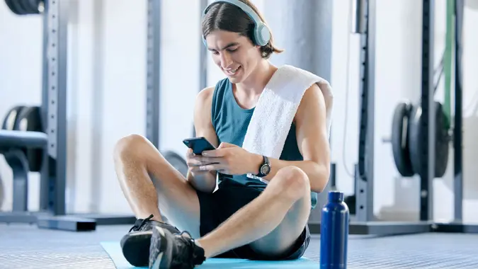 Man check social media in gym, workout music playlist on smartphone and typing online conversation. Happy young athlete sitting on floor, sports health application and fitness lifestyle technology stock photo