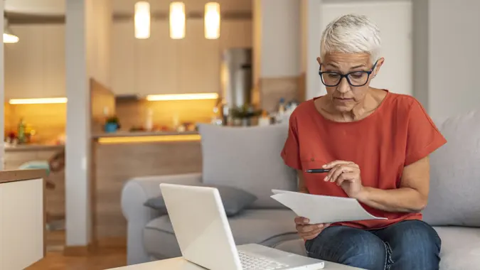 Serious aged woman in eyeglasses checking all bills, calculating expenses.