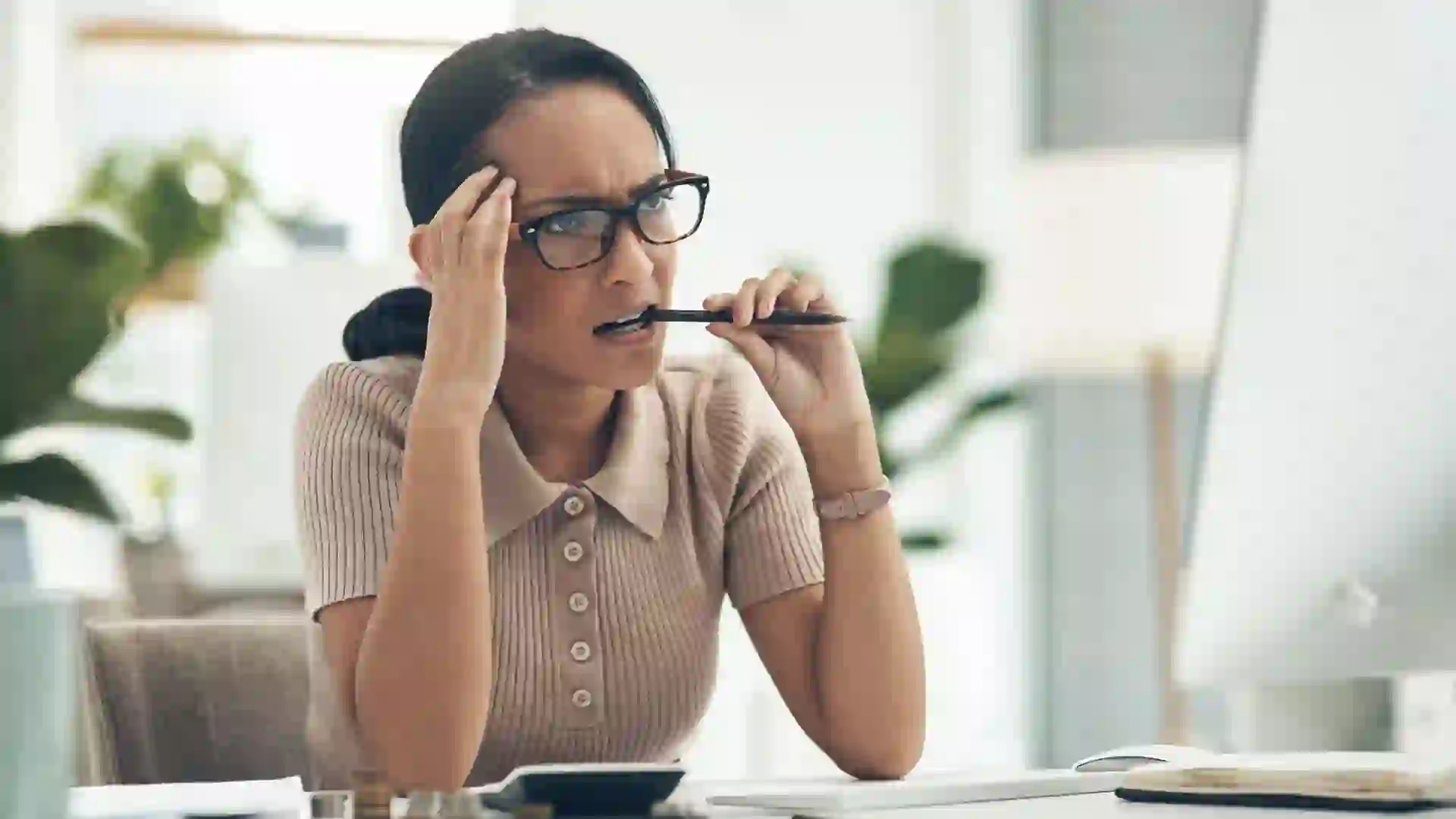 Woman with a worried expression sits at a desk, biting the tip of a pen and looking at a computer.
