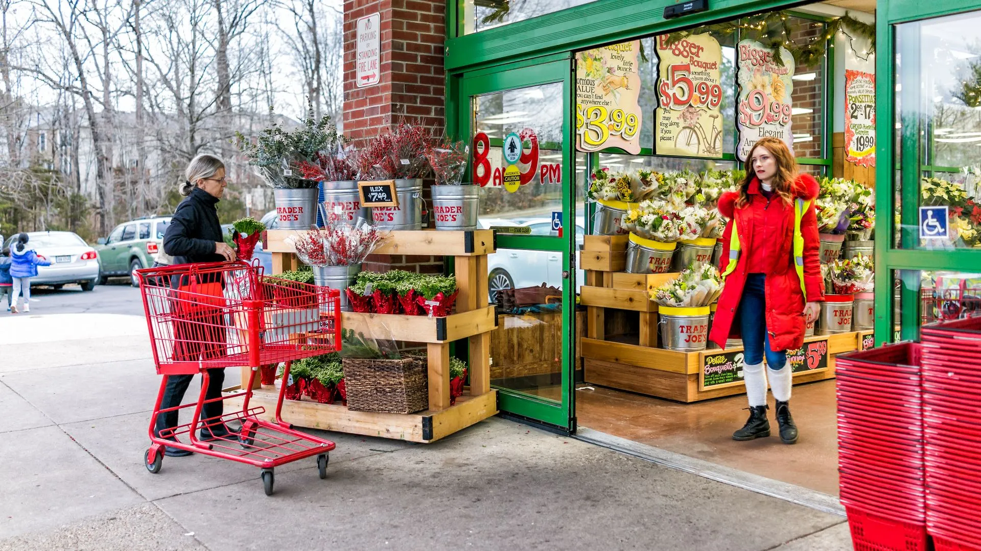 Reston, USA - December 18, 2017: Trader Joe's employee in red clothes with customer, trolley shopping carts by store entrance doors outside women, winter flower pots, gardening plants in Virginia.