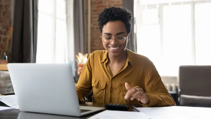Happy young Afro American entrepreneur woman in glasses counting profit, on calculator at laptop computer, analyzing benefits, enjoying financial success, job high result, smiling.