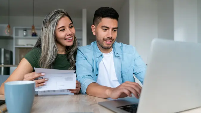 Happy Latin American couple at home paying bills online on their laptop and smiling - financial technology concepts.