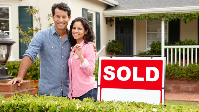 Hispanic couple outside home with sold sign holding keys in hand looking at camera smiling.