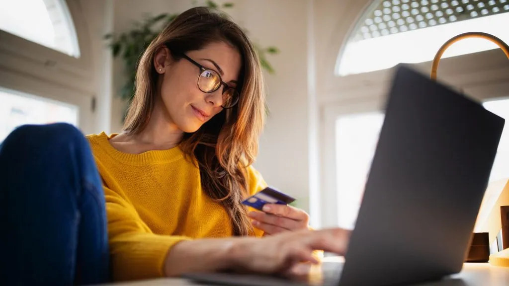 A beautiful young woman enjoying working at home on her laptop in cozy and bright apartment wearing yellow sweater and shopping online paying with credit card.