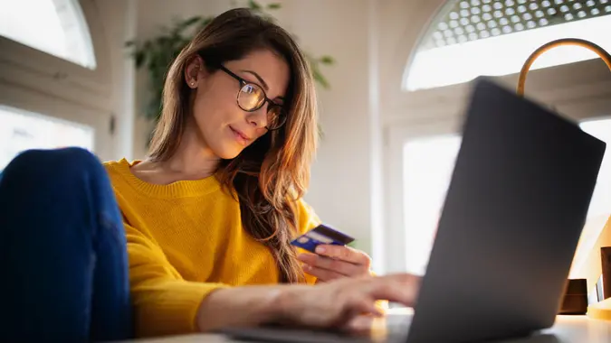 Beautiful young woman in a yellow sweater enjoying working from home on her laptop and shopping online with her credit card in her cozy bright apartment.