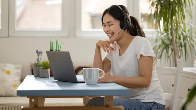Customer support contact center female employee of Mongolian ethnicity working remotely from home, using a laptop, wearing a headset and resolving a customer’s issue with a smile.
