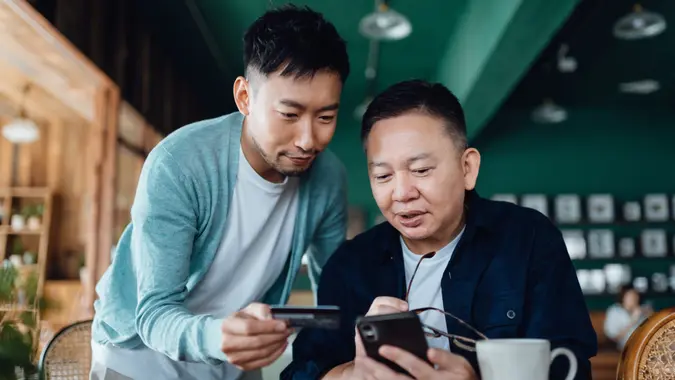 Joyful senior Asian father and son shopping online together using smart phone mobile app and paying online with credit card at home.