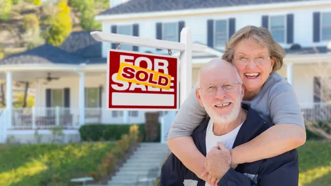 Senior Adult Couple in Front of Sold Home For Sale Real Estate Sign and Beautiful House.