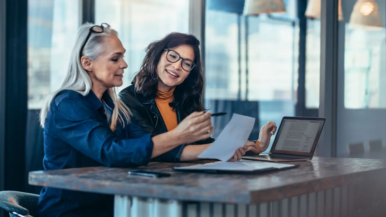 Two women analyzing documents while sitting on a table in office.