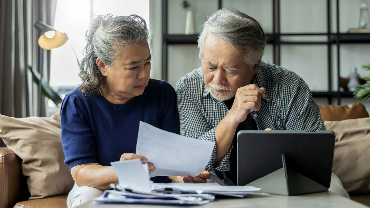 old retired asian senior couple checking and calculate financial billing together on sofa involved in financial paperwork, paying taxes online using e-banking laptop at living room home background.