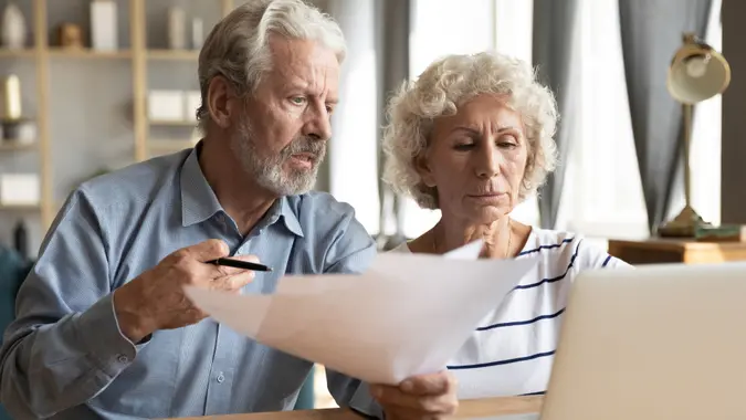 Stressed elderly married couple sitting at table at home manage budget paying using on-line banking access analyzing finance papers, having financial problems, high taxes expenses, unpaid debt concept.