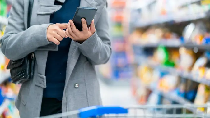 Closeup asian female hand hold smartphone checking price compare in supermarket store with trolley shopping kart with row of products stock photo