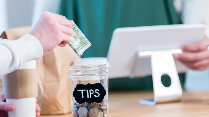 An unrecognizable coffee shop customer stands across the checkout counter from an unrecognizable barista and reaches out to put a paper bill in the tips jar.