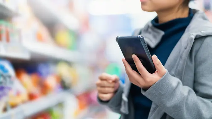 Closeup asian female hand hold smartphone checking price compare in supermarket store with trolley shopping kart with row of products stock photo