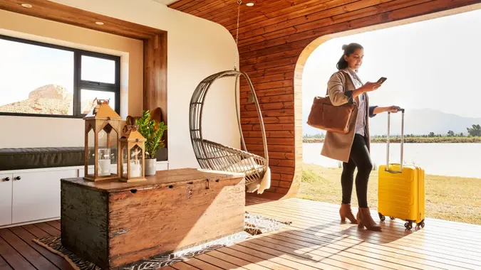Young woman with a suitcase using her mobile phone while standing at lounge of her vacation home during a trip.