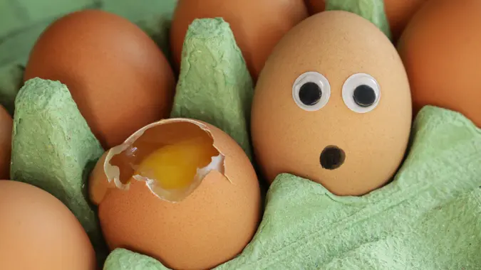 Full frame image of green cardboard, egg box containing batch of chicken eggs, one broken egg with view of yolk and albumen beside egg with googly eye cartoon face expressing shock and horror, focus on foreground stock photo